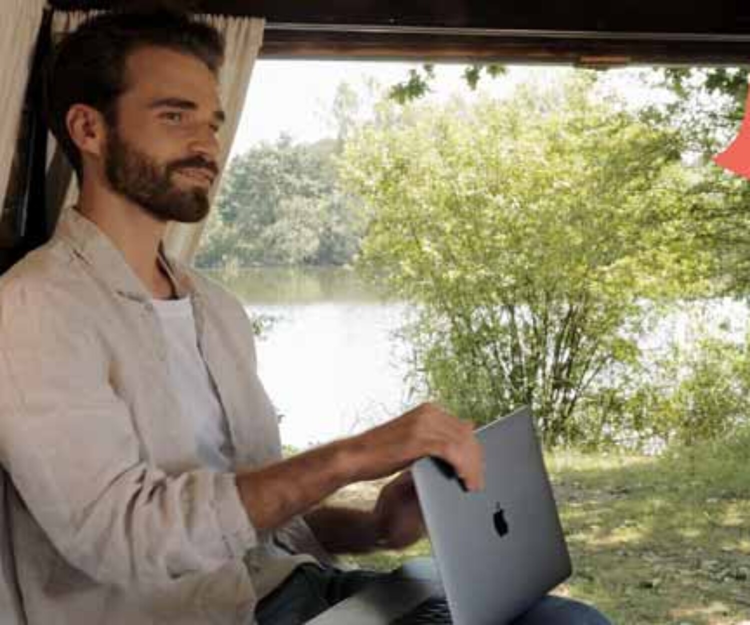 Videoportrait Hannover Junger bärtiger Mann arbeitet mit einem Laptop im Camper mit Blick auf Natur, Bäume und Wasser. Über dem Bild steht „Jetzt bewerben“. Titelbild eines von 52° Film produzierten Recruitingvideos.