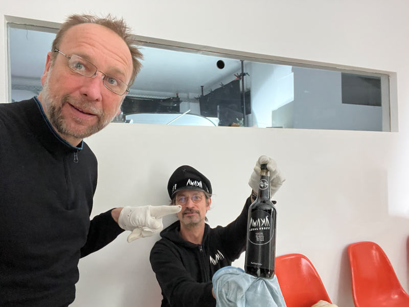 Start 10 Kameramann und DOP Udo Schloermann von der Videoproduktion 52Gradfilm aus Hannover steht vor Papa AWAKA , dieser hält eine Flasche AWAKA hoch und Udo Schloermann zeigt mit einem finger, der in einem Handschuh steckt, auf die Flasche.