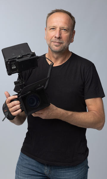 Ueber 52°Film 1 Videograf Hannover Udo Schlörmann steht mit der Filmkamera im Studio vor grauem Hintergrund und lächelt.